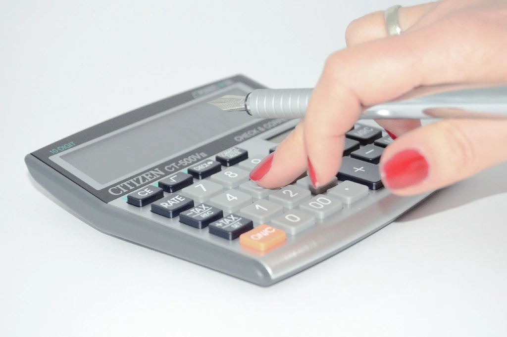 Hand mit rotem Nagellack tippt mit Stift Grunderwerbsteuer-Betrag in einen grauen Taschenrechner ein.