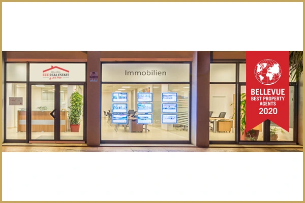 Modernes Immobilienbüro mit großen Fenstern, Leuchtdisplays und "CCC Real Estate"-Schild. Rotes Banner: "BELLEVUE Beste Immobilienmakler 2020".