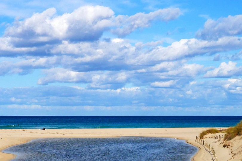 Sandstrand Cala Mesquida mit einer ruhigen Lagune, blauem Meer, weißen Wolken und grasbewachsenen Dünen auf der rechten Seite.