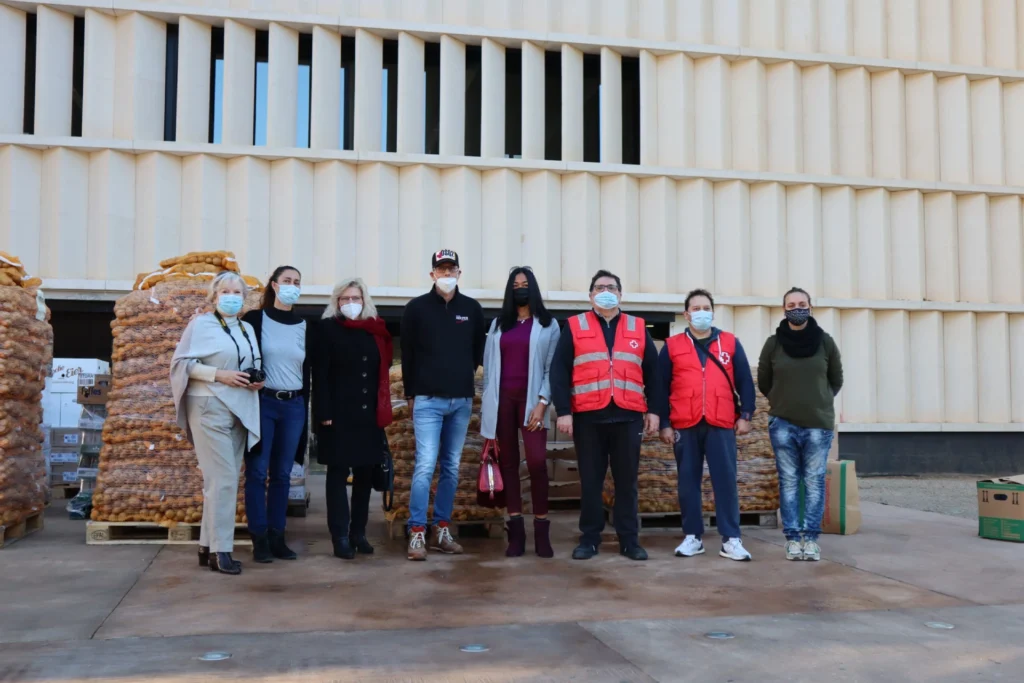 Acht maskierte Personen vor Paletten mit Kartoffelsäcken eines Hilfstransports; zwei tragen rote Westen, beige Gebäude im Hintergrund.
