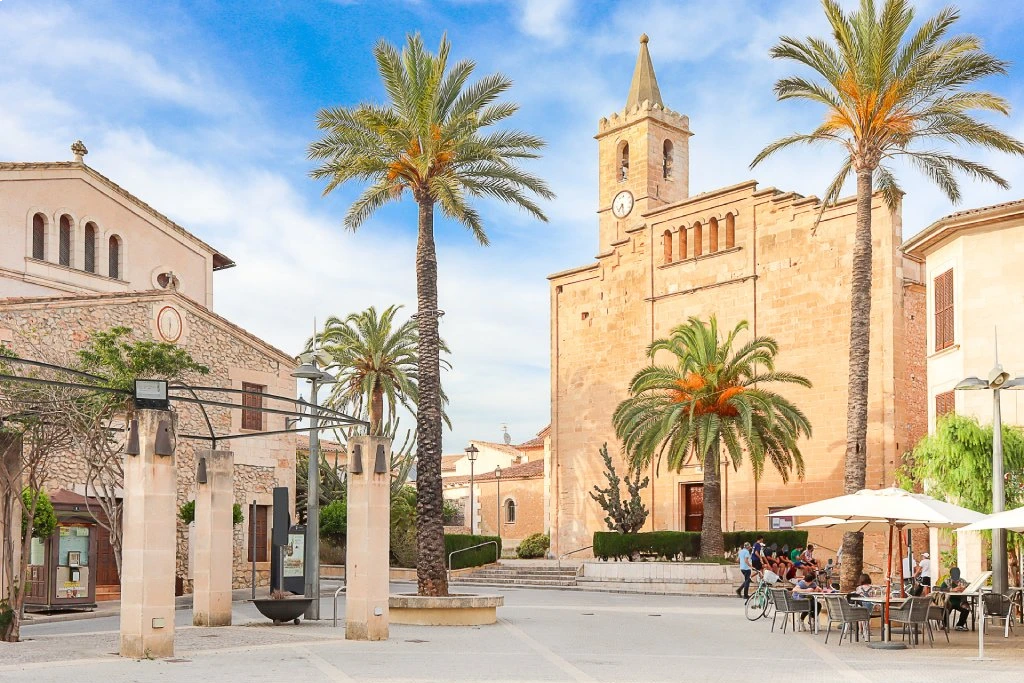 Sonniger Stadtplatz in Sant Llorenç mit Palmen, Außengastronomie und historischer Steinkirche unter blauem Himmel.