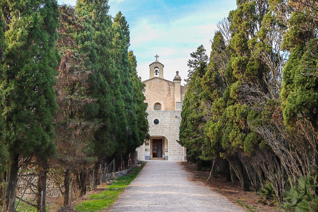 Ein Steinweg mit Zypressen führt unter blauem Himmel zur historischen Kirche Ermita de Betlem.