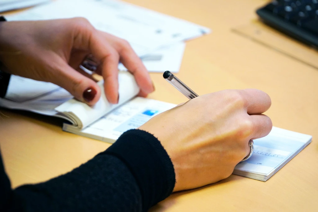 Person schreibt mit Stift einen Scheck, hält ein Scheckheft; im Hintergrund Tastatur und Papiere auf dem Schreibtisch.