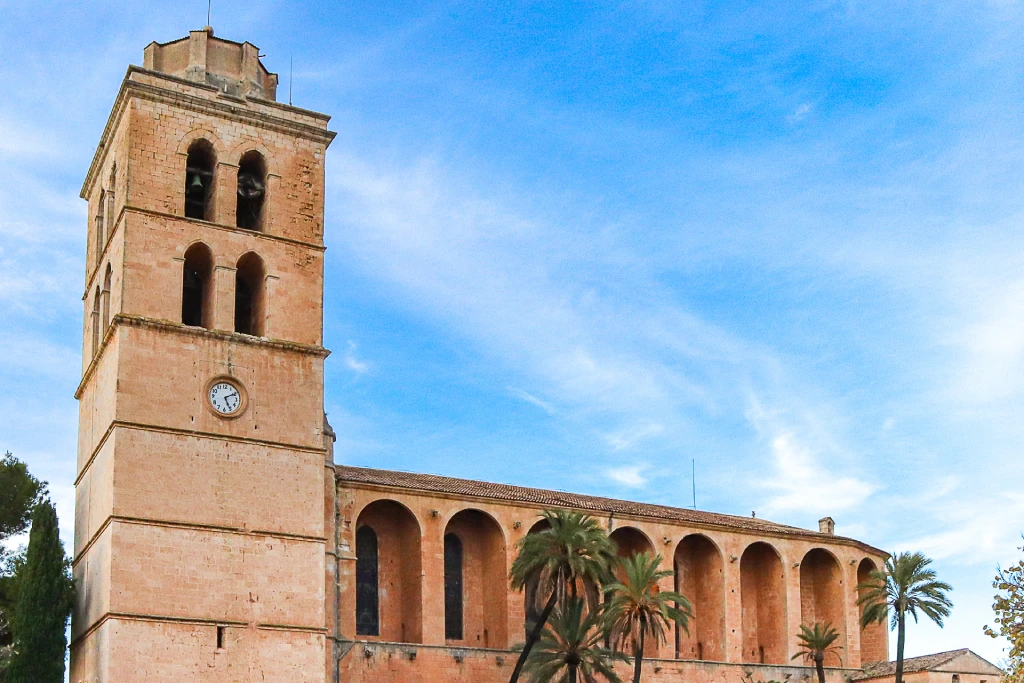 Historische Steinkirche mit Glockenturm und Rundbogenfenstern, umgeben von Palmen unter blauem Himmel in Muro, Mallorca.