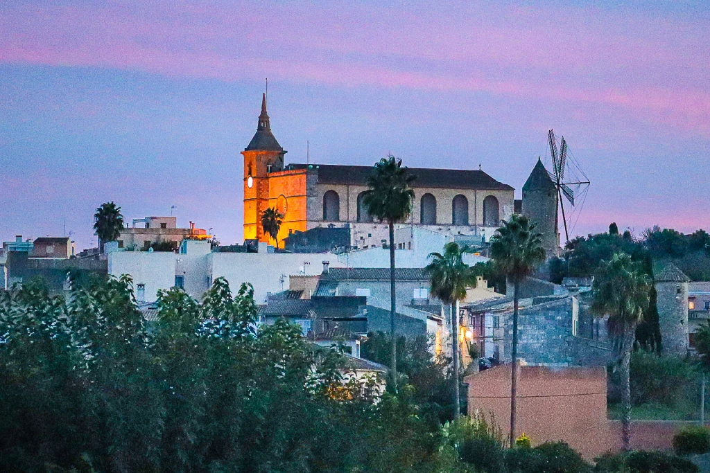 Kirche mit hohem Glockenturm über Santa Margalida in der Abenddämmerung, beleuchtete Mauern, Windmühle, Palmen und violetter Himmel.