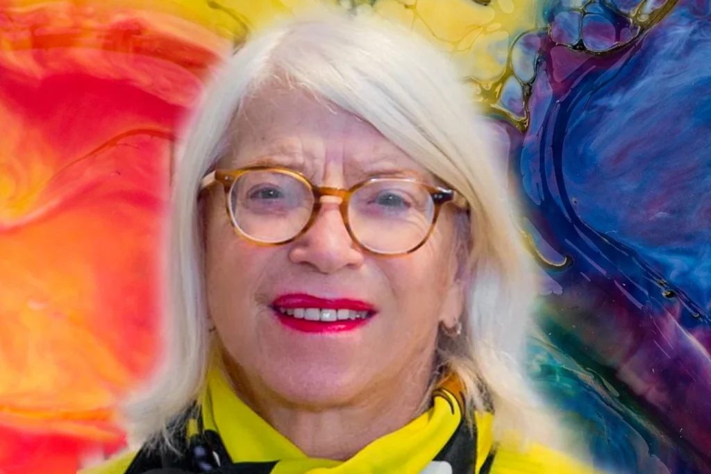 Barbara Radinger, ältere Frau mit weißem Haar, Brille und gelbem Schal, lächelt vor buntem abstraktem Hintergrund.