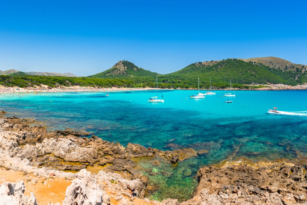 Cala Agulla: Türkisblaues Wasser, Felsen, Segelboote und grüne Hügel unter blauem Himmel im Nordosten Mallorcas.
