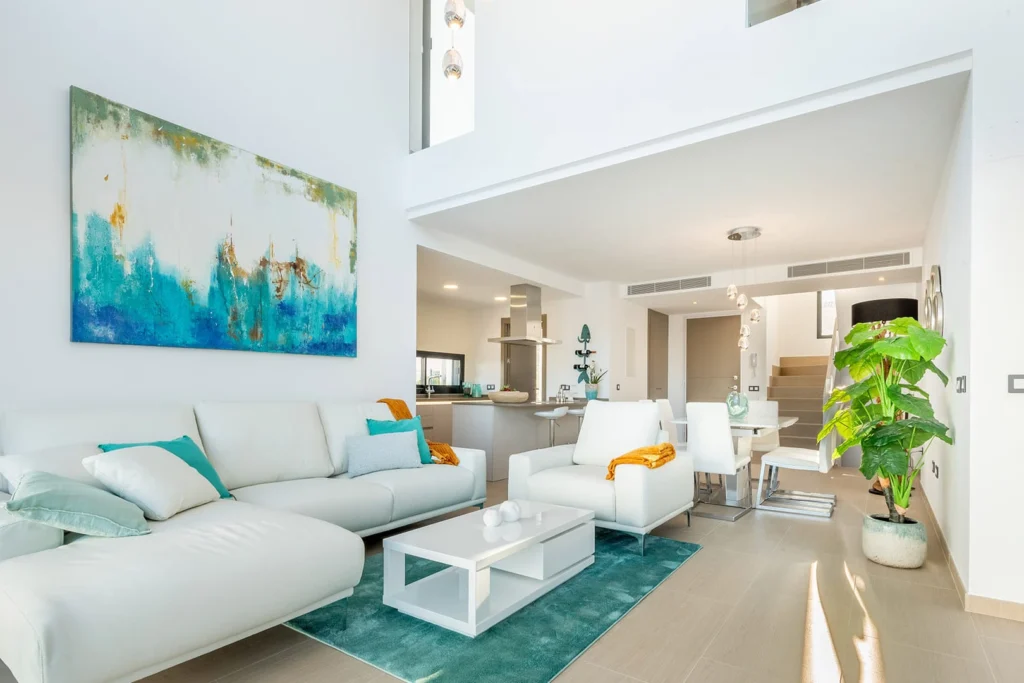 Modernes, helles Wohnzimmer mit weißen Möbeln, türkisen Akzenten und großem Gemälde; offener Grundriss zu Küche und Treppe.