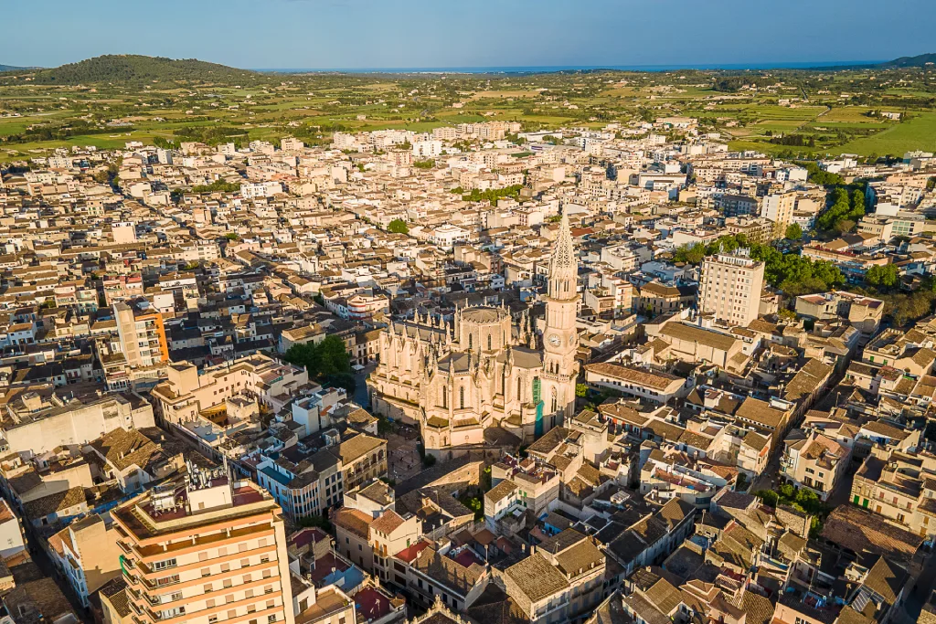Luftaufnahme von Manacor, Spanien: zentrale verzierte Kirche, dichte Bebauung, grüne Felder, entfernte Hügel unter klarem Himmel.
