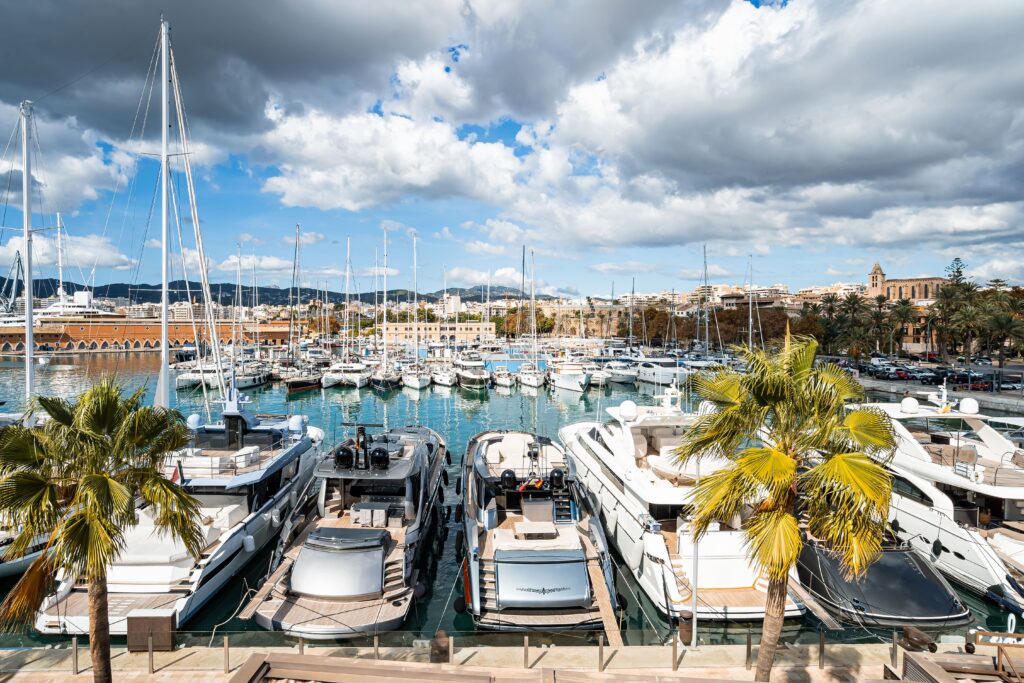 Yachthafen mit Yachten, Segelbooten, Palmen und Gebäuden von Mallorca Immobilien unter einem teilweise bewölkten, sonnenbeschienenen Himmel.