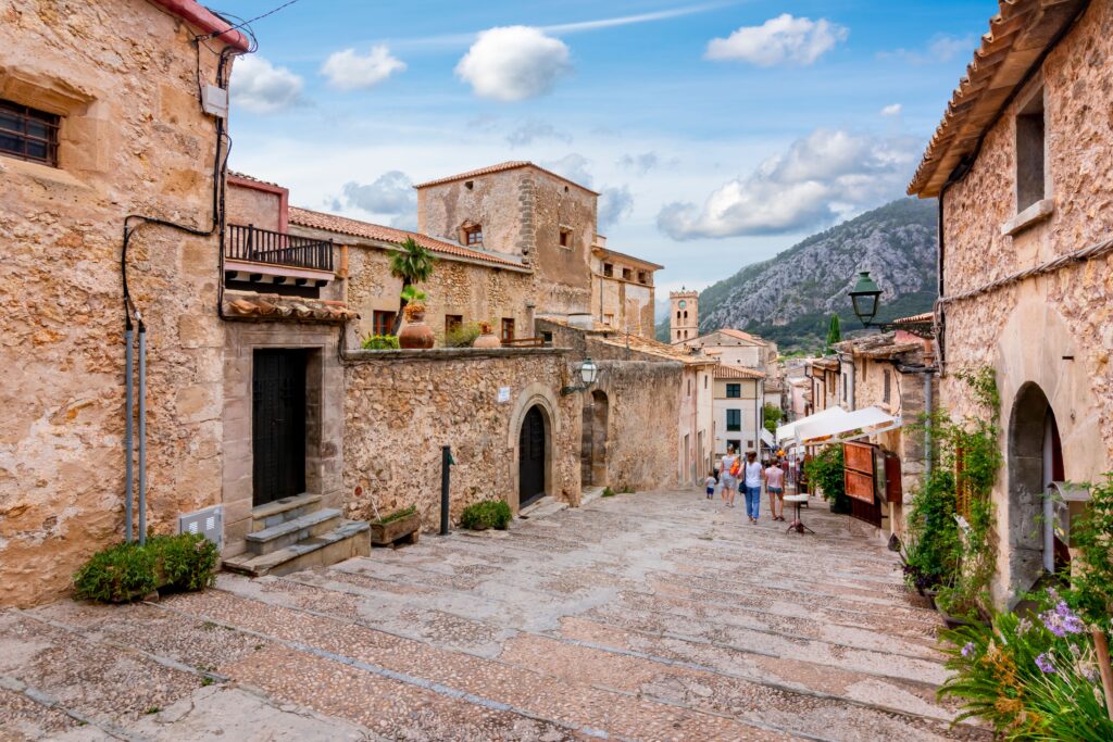 Gepflasterte Straße mit Steinhäusern, Schildern von Mallorca Immobilien, Menschen, die spazieren gehen, Berge und teilweise bewölkter Himmel im Hintergrund.
