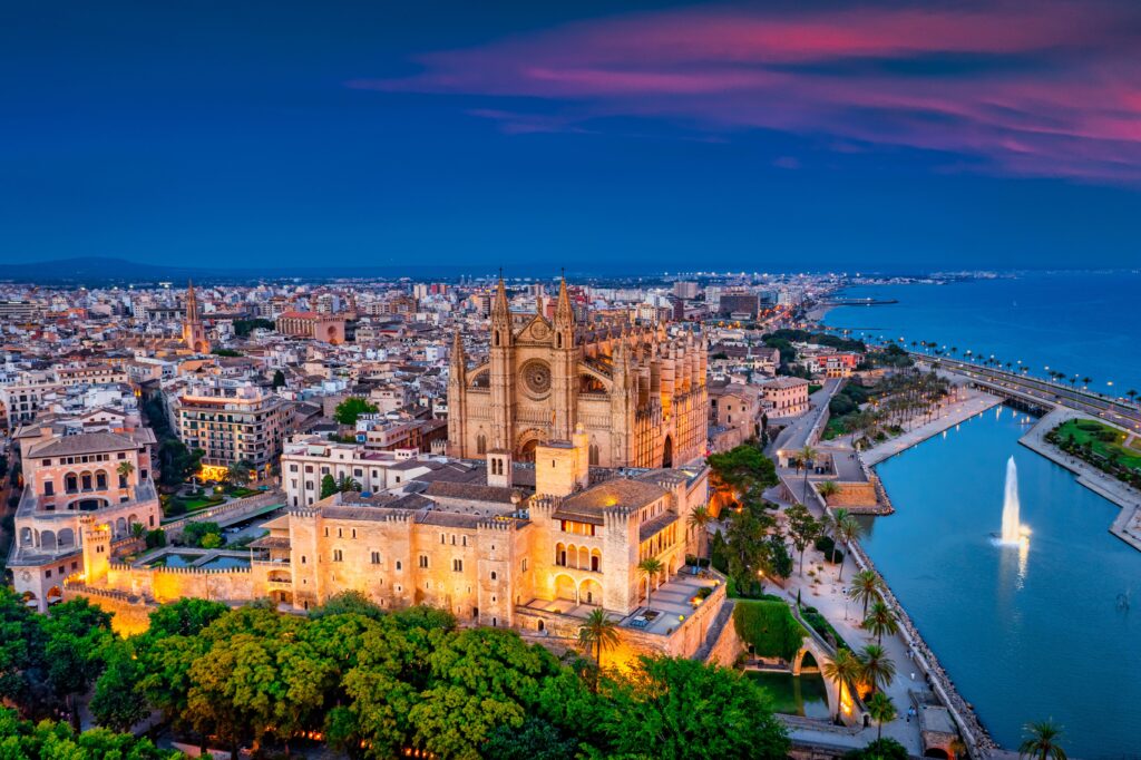 Luftaufnahme von Palma de Mallorca in der Abenddämmerung mit der beleuchteten Kathedrale La Seu, den Lichtern der Stadt und dem ruhigen Meer unter dem bunten Himmel.