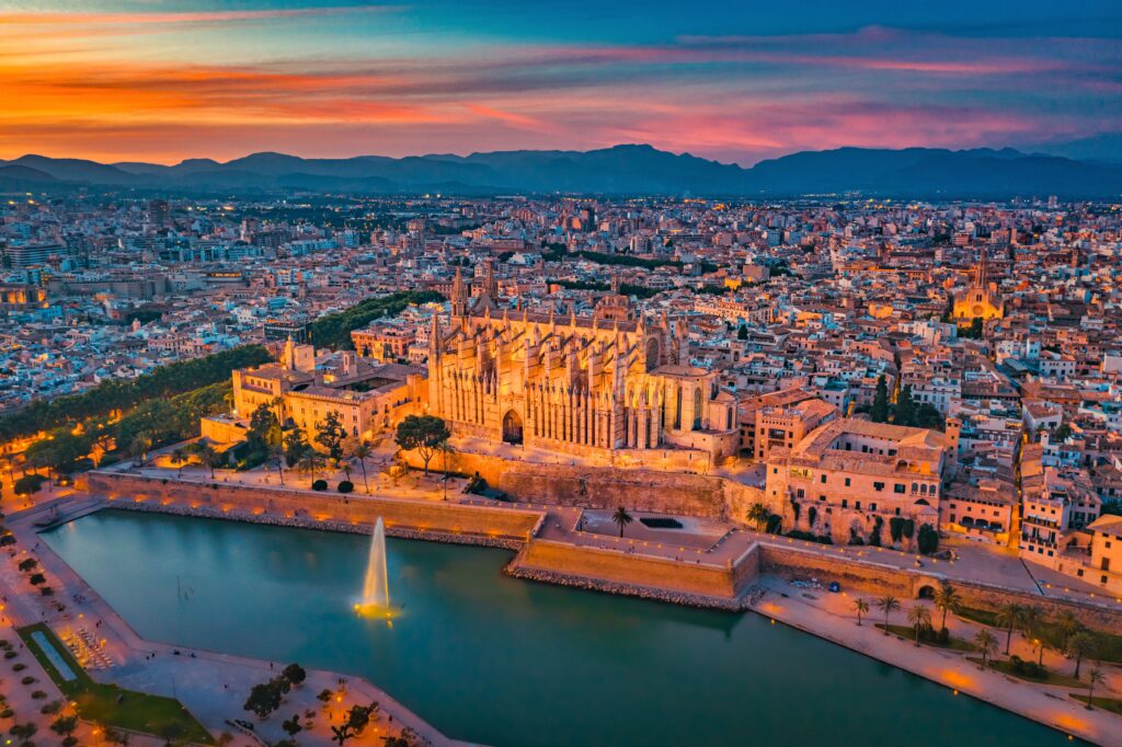 Die Kathedrale von Palma bei Sonnenuntergang auf Mallorca, umgeben von städtischen Gebäuden und einem Springbrunnen am Wasser unter einem strahlenden Himmel.