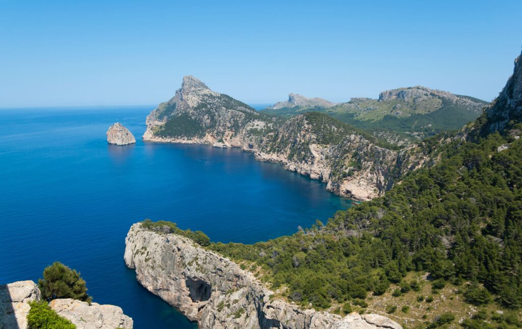 Die zerklüftete Küste Mallorcas mit ihren Steilküsten, grünen Hügeln, dem klaren blauen Meer und den fernen Gipfeln unter einem strahlenden Himmel.
