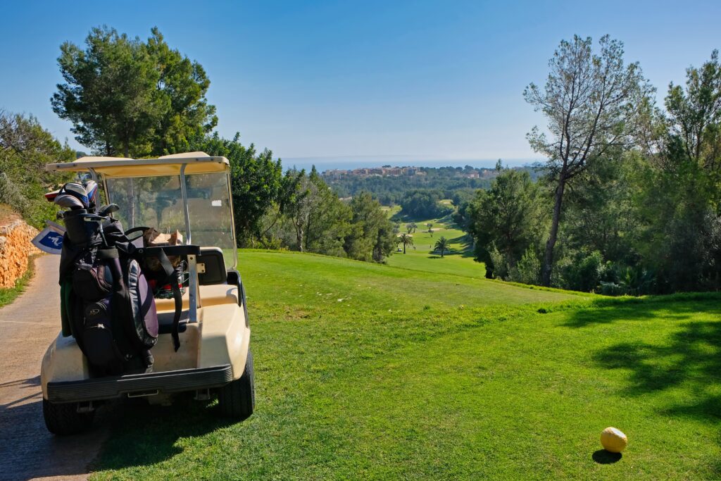 Golfwagen mit Taschen auf dem üppigen Fairway von Mallorca, Bäume rundherum, unter einem klaren blauen Himmel.
