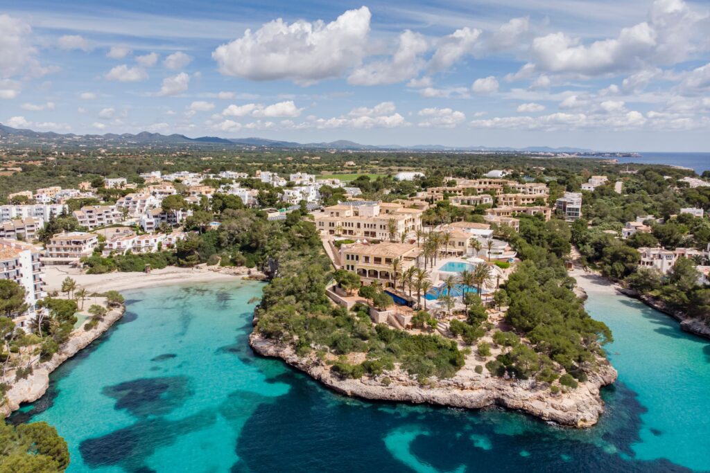 Luftaufnahme des Küstenorts Mallorca mit türkisfarbenem Wasser, Sandstränden, weißen Gebäuden, grünen Bäumen und Bergen.