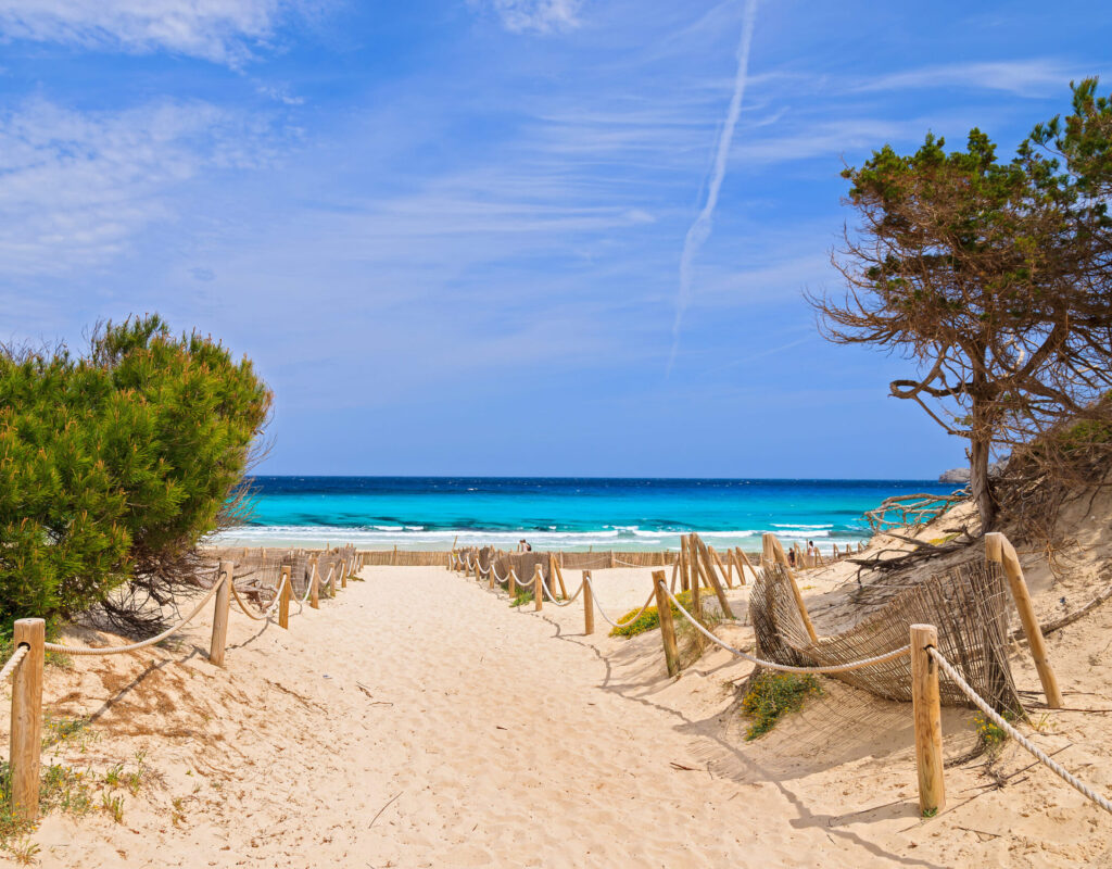 Ein sandiger Weg mit Seilzäunen und Sträuchern führt zum blauen Meer auf Mallorca, unter einem teilweise bewölkten Himmel mit sanften Wellen.