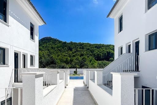 Ein Weg zwischen zwei modernen weißen Gebäuden führt zu einem Pool, mit grünen Bäumen, einem Hügel und blauem Himmel im Hintergrund.