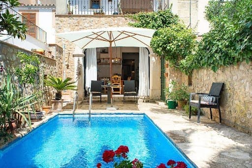 Kleiner Pool mit Steinmauern, viel Grün und Blumen, daneben Essbereich mit weißem Schirm bei offenen Türen zum Haus.