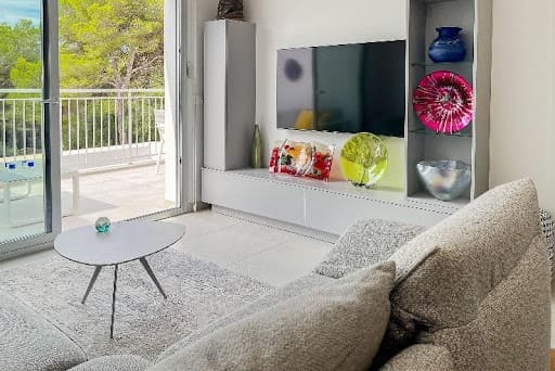 Modernes Wohnzimmer mit grauem Sofa, Couchtisch, Wand-TV, Glasvasen und Balkontüren mit Blick auf Mallorca.