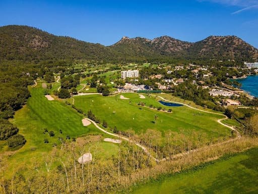 Üppiger Golfplatz mit Bunkern, Wasser, Bäumen, weit entfernten Gebäuden und Küstenlinie unter einem klaren blauen Himmel auf Mallorca.