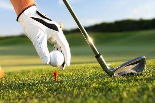 Golfer mit weißem Handschuh setzt Ball auf roten Tee, Golfschläger daneben, sonniger Rasen auf einem Golfplatz Mallorca.