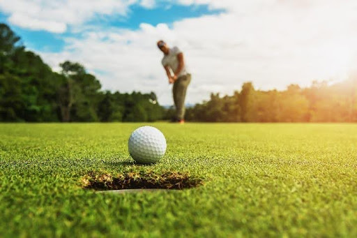 Golfball am Lochrand auf mallorquinischem Grün, Golfer im Hintergrund beim Putten an einem sonnigen Tag.