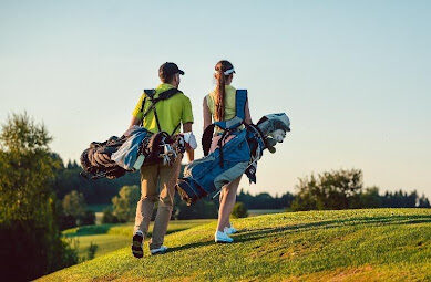 Zwei Golfer mit Taschen gehen bei Sonnenschein einen Hügel hoch, umgeben von Bäumen und weitem Himmel auf Mallorca.